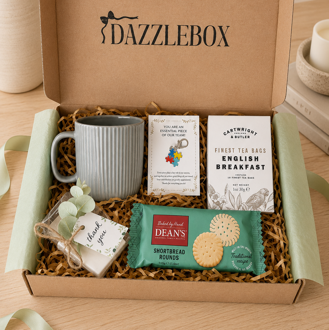 A DazzleBox gourmet gift box set on a wooden table, containing a gray ribbed ceramic mug, a box of Earl Grey English Breakfast tea, a pack of Dean's Shortbread Biscuits, a small 'Thank You' card in a ribbon loop, and shredded paper filler.