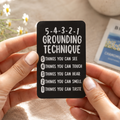 A person holds a black rectangular card with white text reading '5-4-3-2-1 GROUNDING TECHNIQUE' and a numbered list. The scene is softly lit with a beige fabric background, small daisy flowers, a ceramic cup, and a Birchall chamomile flower packet nearby.