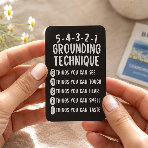 A person holds a black rectangular card with white text reading '5-4-3-2-1 GROUNDING TECHNIQUE' and a numbered list. The scene is softly lit with a beige fabric background, small daisy flowers, a ceramic cup, and a Birchall chamomile flower packet nearby.