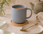 A ribbed ceramic mug filled with a light brown beverage sits on a round wooden coaster on a beige tablecloth. A matching beige plate with a gold spoon rests nearby, with a ceramic teapot in the background and soft floral decor in the foreground.