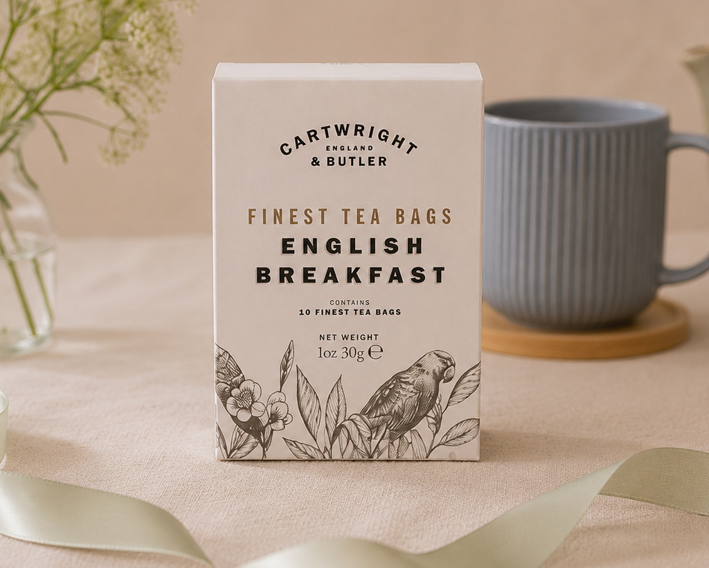 A box of Cartwright & Butler English Breakfast tea stands on a beige fabric surface, styled with a blue-gray mug, flowers in a glass vase, and a ribbon in the background.