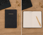 A black hardcover notebook with gold lettering and a matching gold pen, shown in a split view: closed on the left and open on the right on a wooden desk with a laptop, binder clips, and a gold pen.