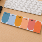 A weekly planner notepad with rounded arches in blue, light blue, orange, peach, coral, and orange, laid out on a light wood desk next to a keyboard, notebook, and pen.