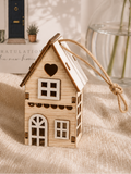 A small wooden house ornament with a hanging string sits on a beige fabric surface. Behind it is a greeting card reading 'Congratulations on the new home,' with baby's breath flowers in a glass vase in the background.