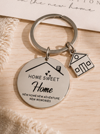 A round engraved metal keychain with a house charm and a small house charm, displayed on a light wood surface next to a congratulatory card for a new home.