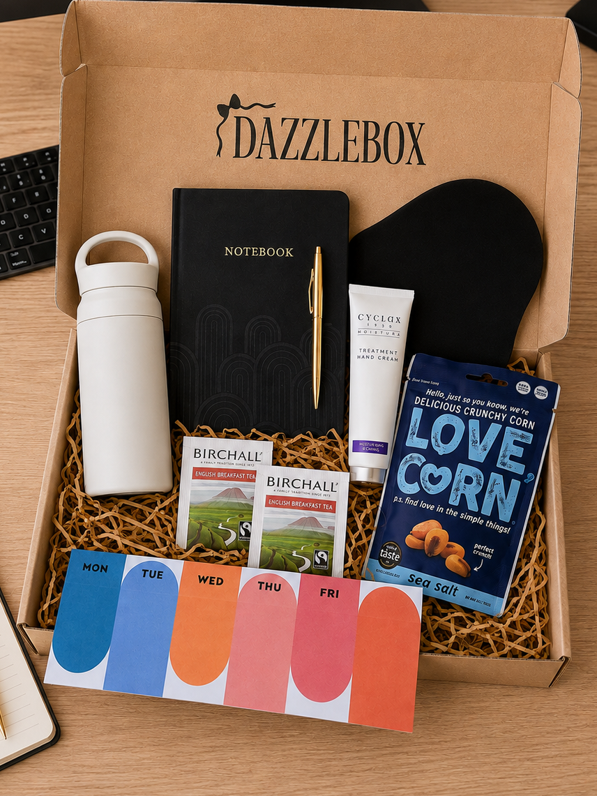 An open Dazzlebox gift box on a wooden desk, filled with curated items including a black notebook, white tumbler, black headband, tube of lip balm, two Birchfield tea boxes, a Love Corn candy package, and a weekly color card set. An Apple iMac is partially visible behind the box.