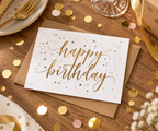 A white 'happy birthday' greeting card with gold script lettering and scattered gold, rose gold, and silver confetti, resting on a brown kraft envelope on a wooden table surrounded by party decorations including a slice of cake, gold ribbon, glass, candle, dazzlebox gift box.