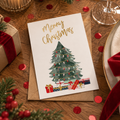 A Christmas greeting card with a watercolor green tree and gold 'Merry Christmas' text, placed on a kraft envelope on a wooden table with candles, dazzlebox gifts, pine branches, and red confetti.