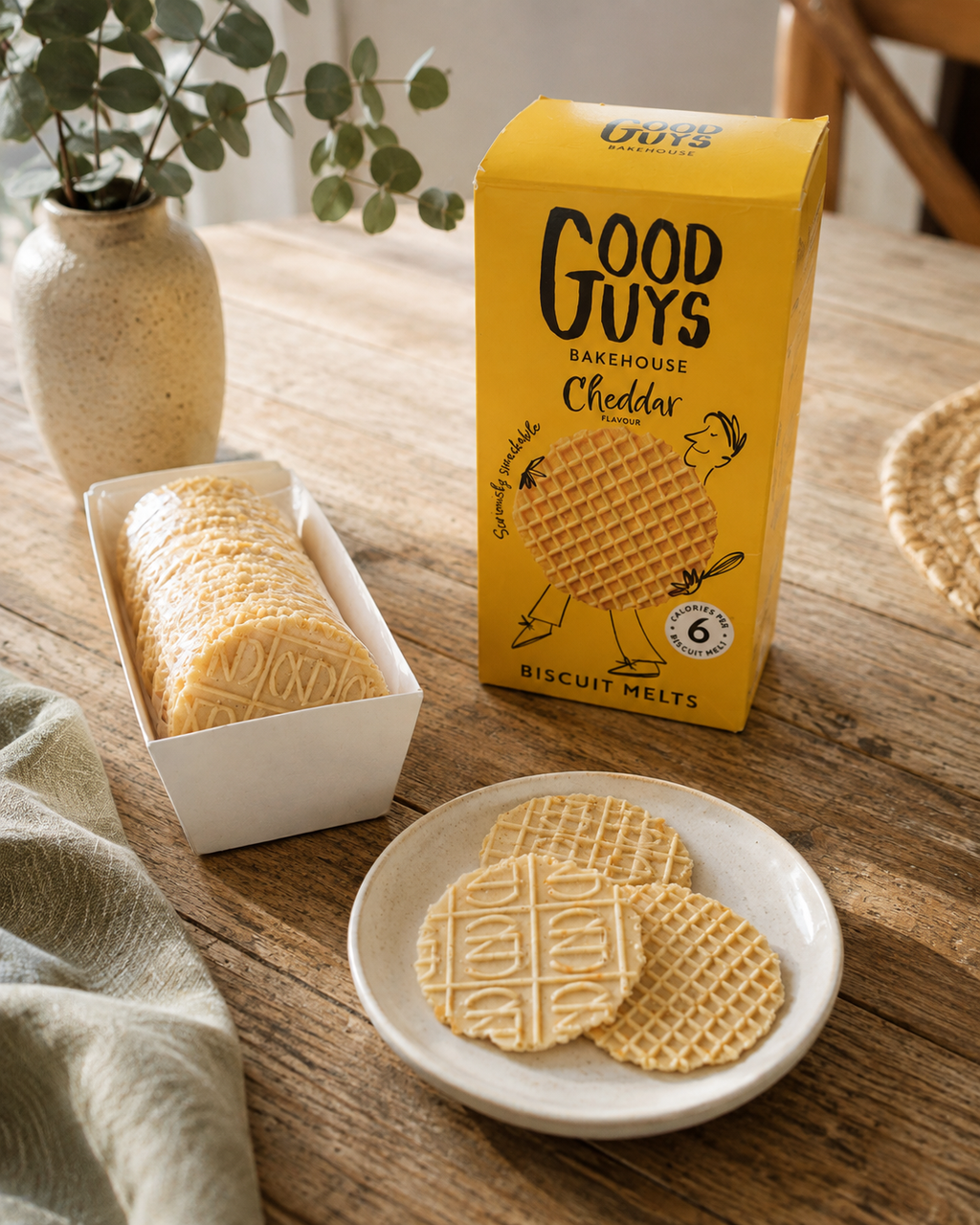 A yellow box of Good Guys Bakehouse Biscuit Melts Cheddar is shown on a wooden table next to a plate of baked biscuit melts and a loaf in a white tray, with a vase of greenery in the background.