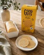 A yellow box of Good Guys Bakehouse Biscuit Melts Cheddar is shown on a wooden table next to a plate of baked biscuit melts and a loaf in a white tray, with a vase of greenery in the background.