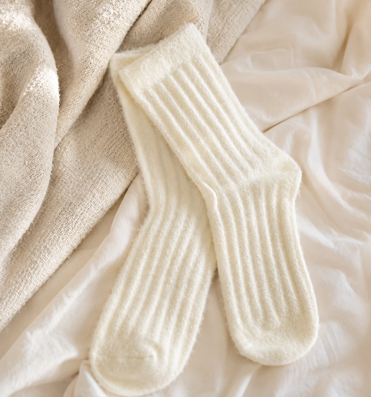 A pair of cream-colored ribbed knit socks laid flat on a soft beige blanket and white fabric background.