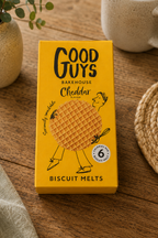 A yellow box of Good Guys Bakehouse Cheddar Biscuit Melts sits on a wooden table, surrounded by a ceramic mug, a small potted plant, and a woven placemat.