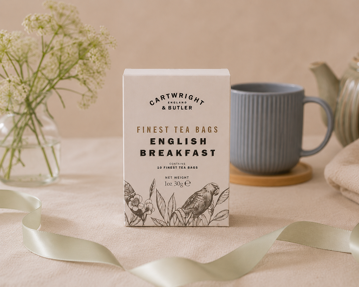 A box of Cartwright & Butler English Breakfast tea stands on a beige fabric surface, styled with a blue-gray mug, flowers in a glass vase, and a ribbon in the background. dazzlebox