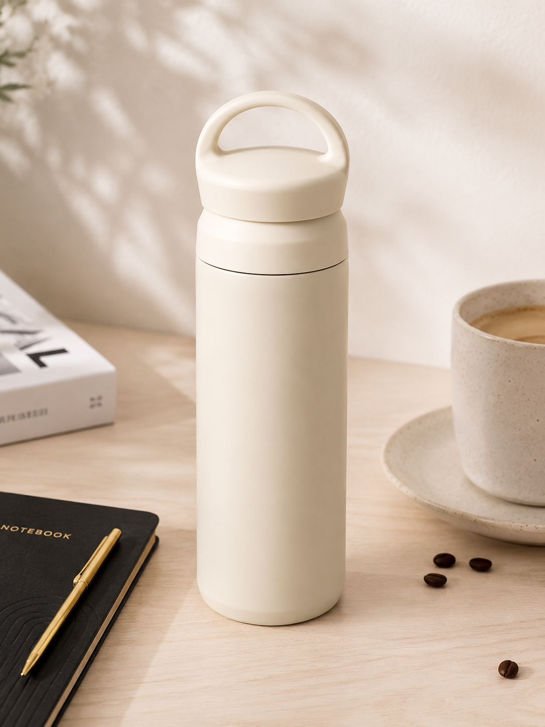 A matte cream-colored insulated water bottle with a matching lid handle, set on a light wood desk next to a notebook, pen, coffee beans, and a cup of coffee.