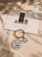 A round engraved metal keychain with a house charm and a small house charm, displayed on a light wood surface next to a congratulatory card for a new home.