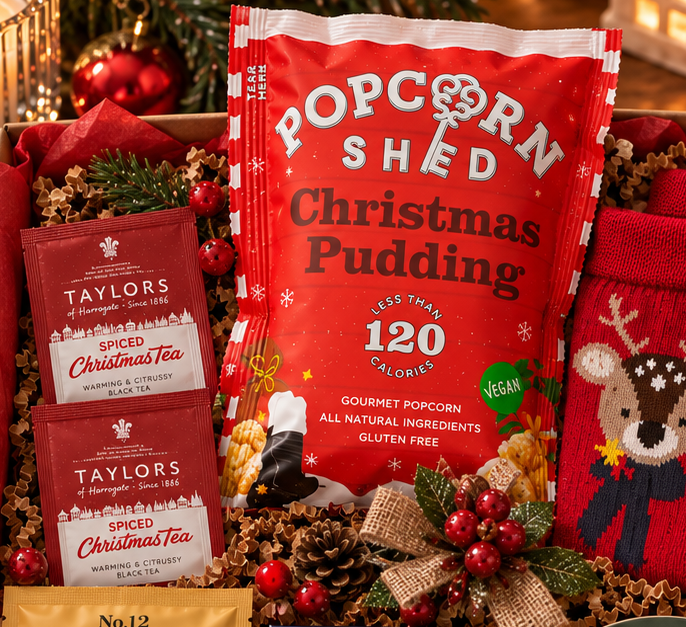 A Christmas Letterbox gift box by dazzlebox filled with holiday treats and cozy items, including a bag of Popcorn Shred Christmas Pudding, Taylors of Harrogate Christmas Tea, Willes Cakes Milk and Gods, a red Christmas-themed sock, a green lip balm tube, a small Christmas plate, and a gold spoon, arranged in a cardboard box with red tissue paper and pine cones. The box is decorated with a 'Merry Christmas' card and surrounded by festive lights, ornaments, and a mug on a wooden table.