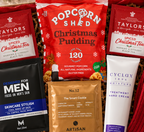 A curated DazzleBox Christmas gift box filled with assorted treats and self-care items, including Taylor's Christmas Tea, popcorn, Skinceutious Men skincare, Artisan Chocolate, Cyclax hand cream, a red knit scarf, and a Christmas card, arranged in a cardboard box with red tissue paper.