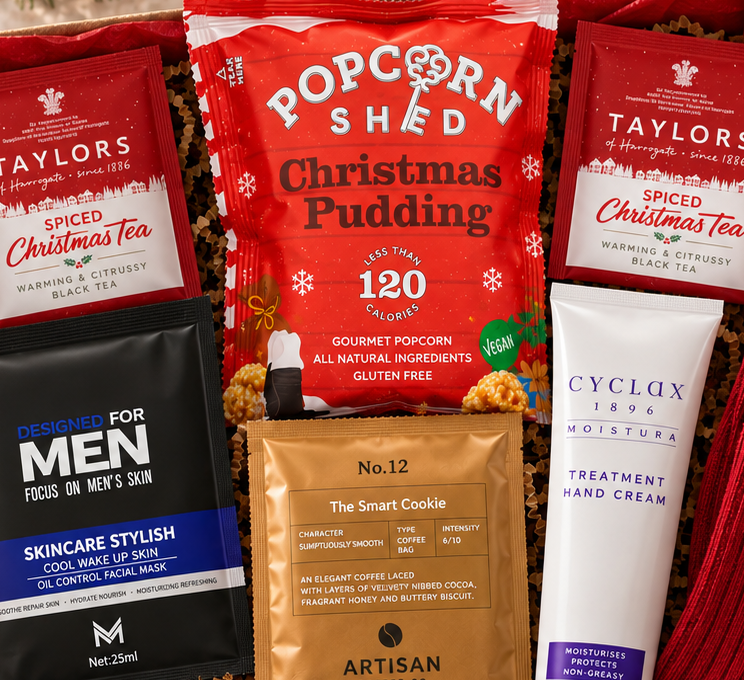 A curated DazzleBox Christmas gift box filled with assorted treats and self-care items, including Taylor's Christmas Tea, popcorn, Skinceutious Men skincare, Artisan Chocolate, Cyclax hand cream, a red knit scarf, and a Christmas card, arranged in a cardboard box with red tissue paper.