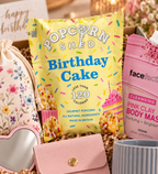 A curated dazzlebox birthday gift box featuring a yellow bag of Popcorn Shed Birthday Cake gourmet popcorn, a pink facefact Pink Clay Body Mask packet, a pink wallet, a floral drawstring pouch with a heart-shaped mirror, and a striped mug, arranged in crinkle paper.