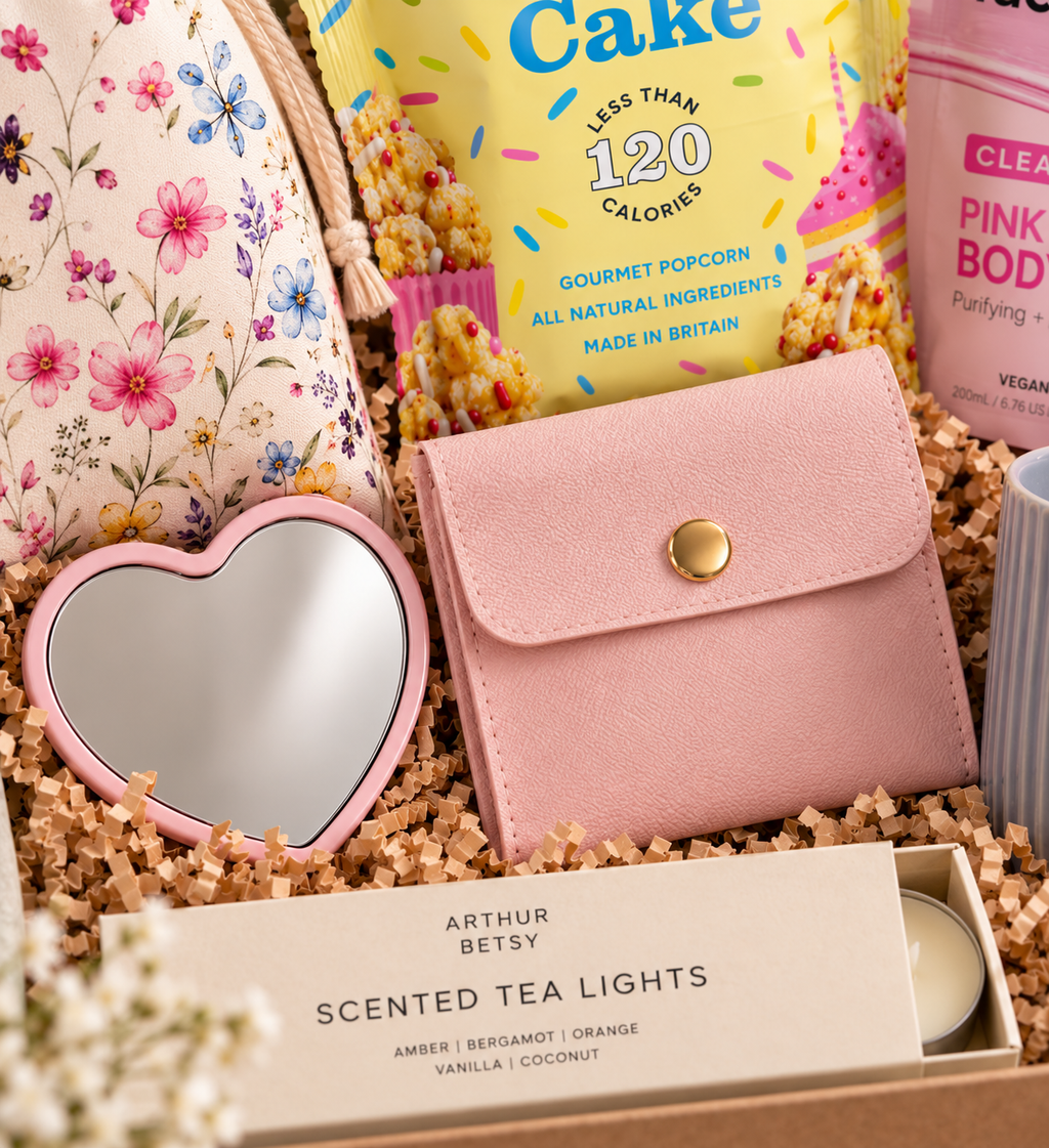 A curated dazzlebox birthday gift box arrangement featuring a pink faux-leather wallet with a gold snap, a heart-shaped pink mirror, a floral fabric pouch, a bag of gourmet popcorn, a pink body wash bottle, and a box of Arthur Betsy Scented Tea Lights nestled in brown paper packing.