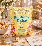 A bag of Popcorn Shed Birthday Cake gourmet popcorn stands on a decorated tabletop with cake slices, popcorn bowls, gold confetti, and a Happy Birthday card. The packaging is bright yellow with blue text and colorful sprinkle graphics. dazzlebox