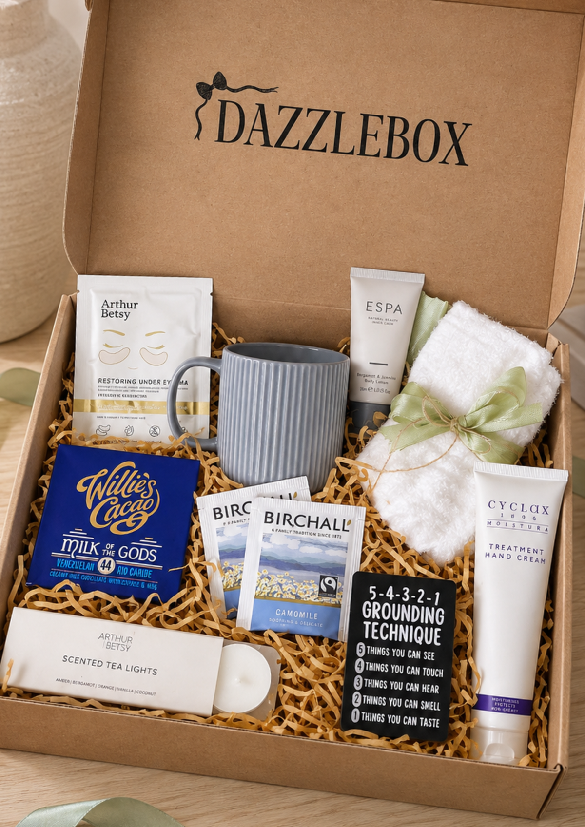 Corporate Wellbeing Gift Box – Employee Wellness & Care Package An open DazzleBox gift box filled with assorted self-care and wellness items, including a gray ceramic mug, hand cream, bath salts, a towel, scented tea lights, and small informational cards, photographed on a light wood surface.