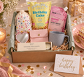 A curated Dazzlebox Birthday gift box for her set styled on a table with a cake slice, flowers, candles, and confetti. The box contains a floral drawstring pouch, a pink body mask packet, a yellow popcorn shed birthday cake bag, a blue-gray mug, a heart-shaped mirror, a scented tea lights box, and a small pink pouch, all arranged in kraft paper filler with a green tissue lining.