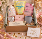 A curated Dazzlebox Birthday gift box for her set styled on a table with a cake slice, flowers, candles, and confetti. The box contains a floral drawstring pouch, a pink body mask packet, a yellow popcorn shed birthday cake bag, a blue-gray mug, a heart-shaped mirror, a scented tea lights box, and a small pink pouch, all arranged in kraft paper filler with a green tissue lining.
