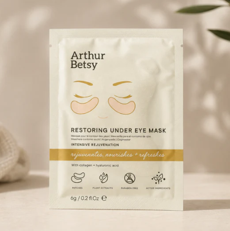 Arthur Betsy Restoring Under Eye Mask sachet in a white packet with gold and black text, featuring a simple eye illustration, set against a softly blurred warm beige background with leaf shadows.