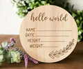 A round natural wood baby shower sign engraved with 'hello world' and fields for name, date, height, and weight, styled on a wooden surface with green foliage and purple flowers in the background.