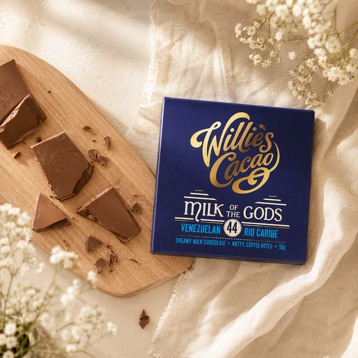 A square blue box of Willie's Cacao Milk of the Gods chocolate is styled on a cream fabric background with a wooden board, broken chocolate pieces, and white baby's breath flowers.