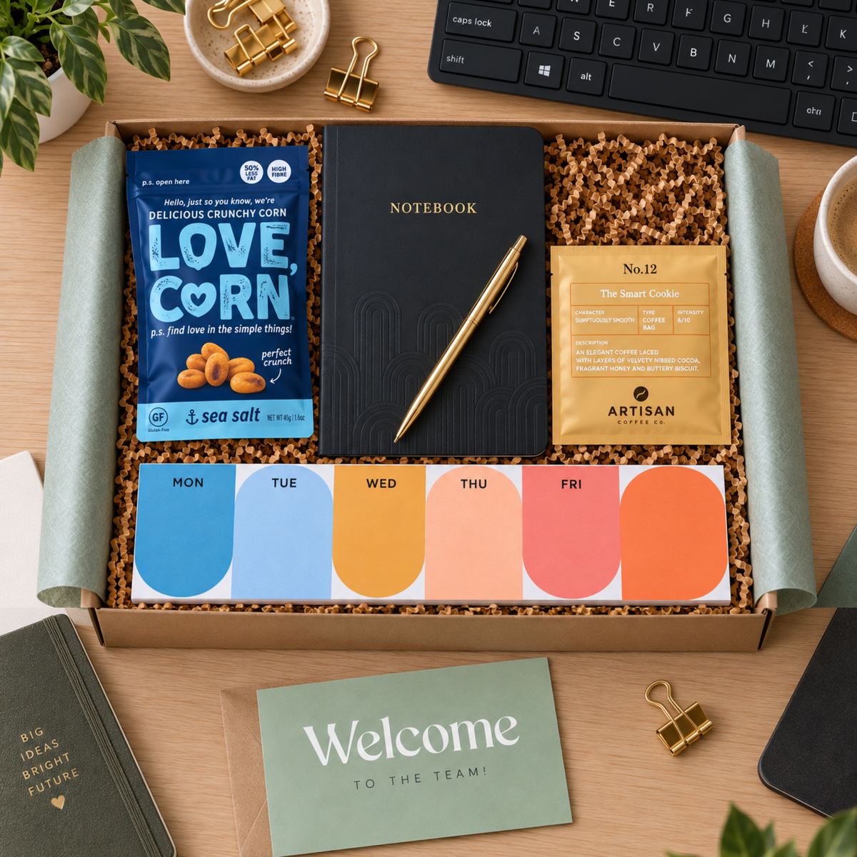 A curated dazzlebox gift box new starter corporate employee gift set displayed on a wooden desk, containing a black notebook with a gold pen, a blue bag of sea salt corn chips labeled 'LOVE CORN', a gold pouch from Artisan Allies, a weekly color card set, and a green 'Welcome to the team!' card.