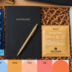A curated dazzlebox gift box letterbox new starter corporate employee gift set gift box set displayed on a wooden desk, containing a black notebook with a gold pen, a blue bag of sea salt corn chips labeled 'LOVE CORN', a gold pouch from Artisan Allies, a weekly color card set, and a green 'Welcome to the team!' card.