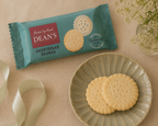 A package of Dean's Shortbread Rounds cookies lies on a beige surface beside a pale green plate holding two round buttery cookies, with a ribbon and small flowers as styled props.