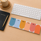 A weekly planner notepad with rounded arches in blue, light blue, orange, peach, coral, and orange, laid out on a light wood desk next to a keyboard, notebook, and pen.